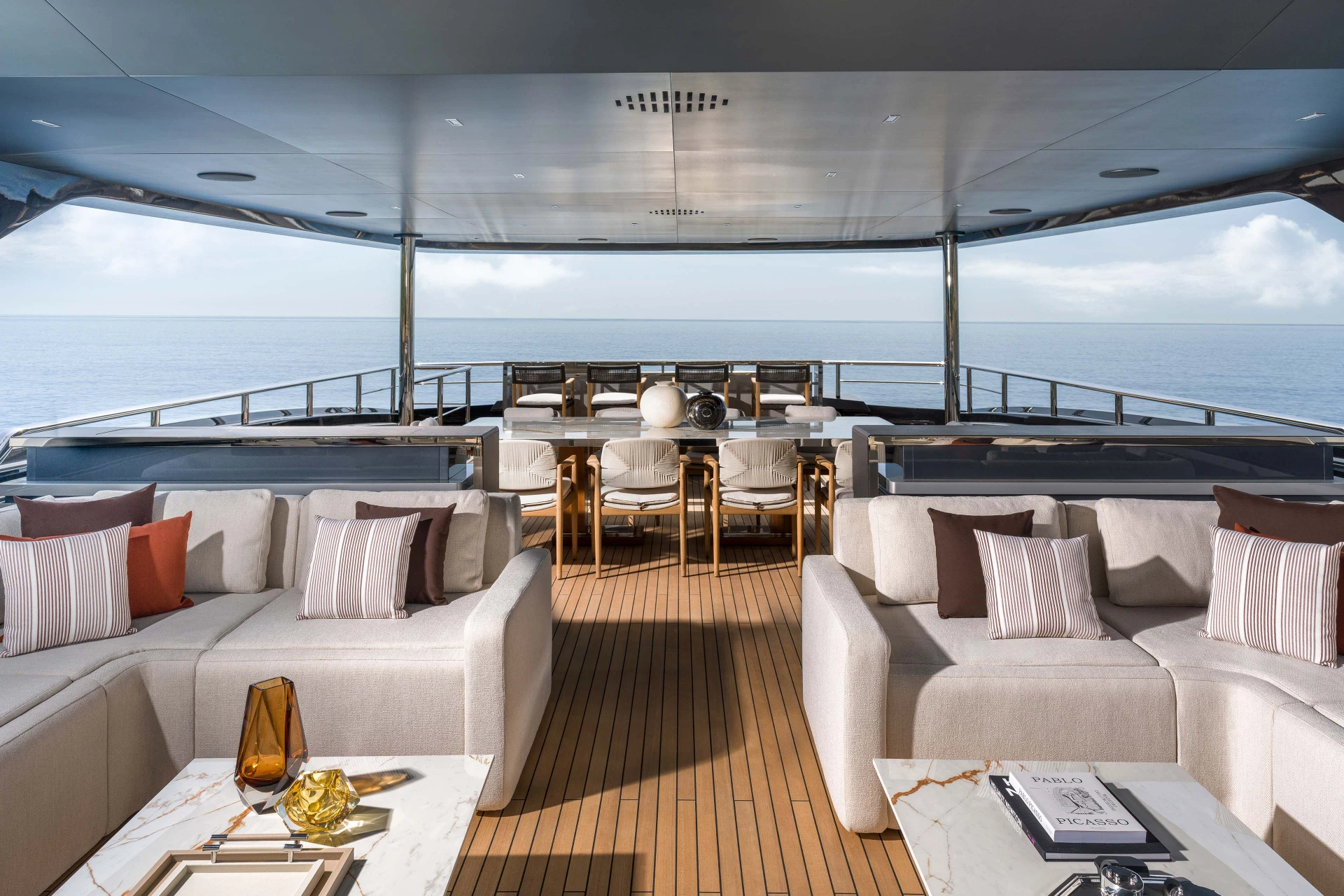 A covered aft deck looking astern over facing sofas to a dining table, with open sea beyond