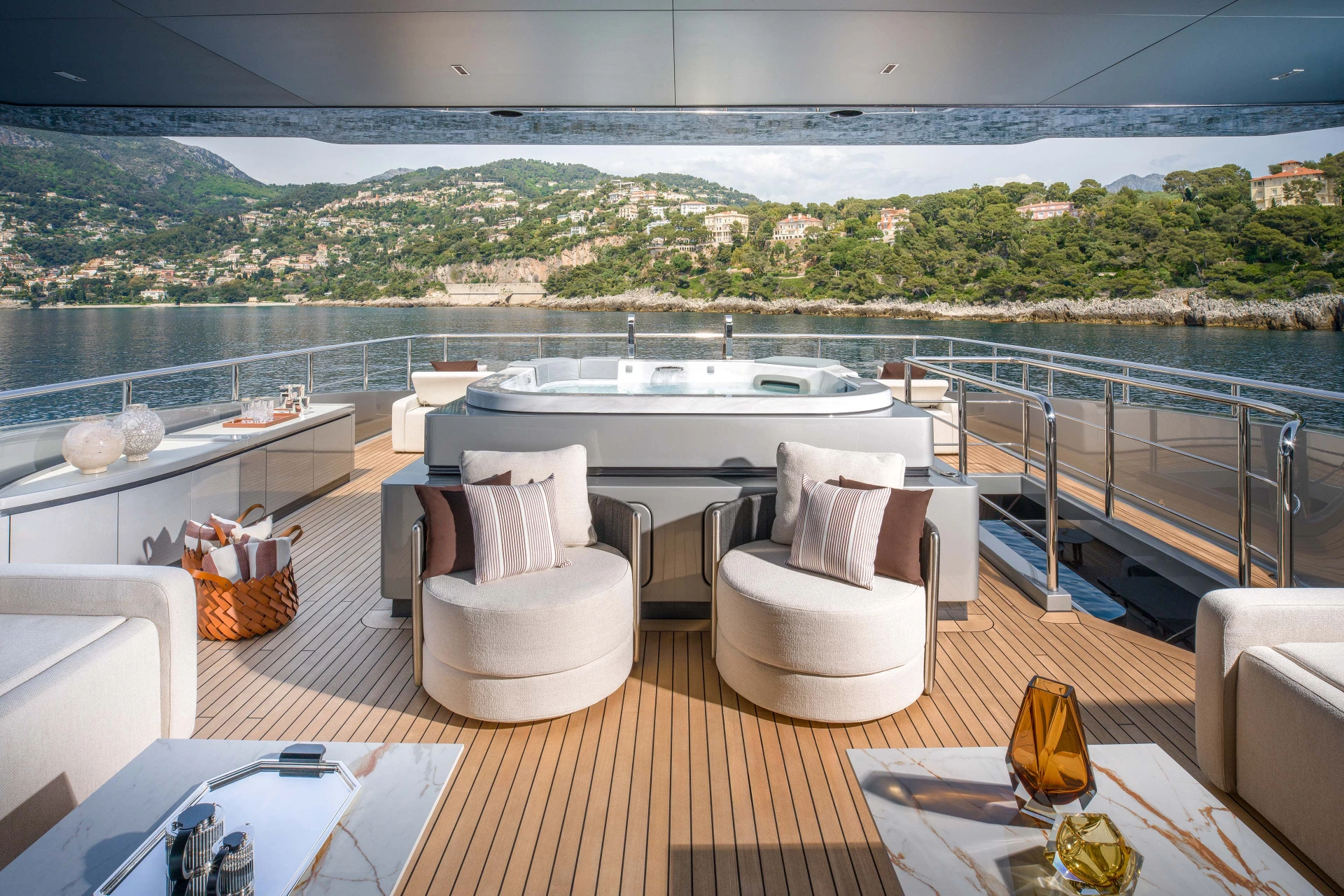 An open deck with a spa pool set into teak, swivel armchairs around it and the Riviera coastline beyond