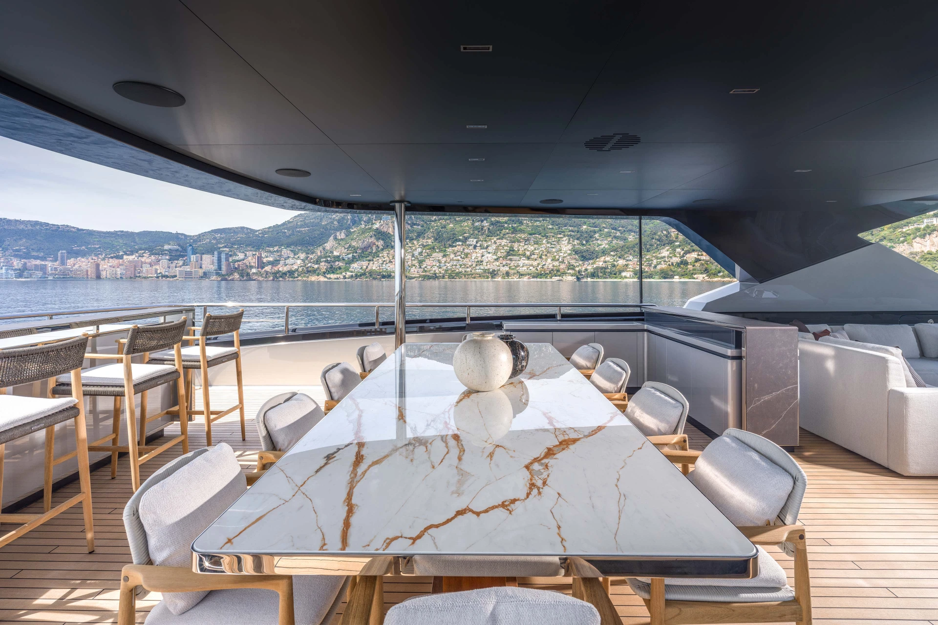 A shaded deck dining table in veined marble with woven bar stools alongside and the Monte Carlo skyline across the water
