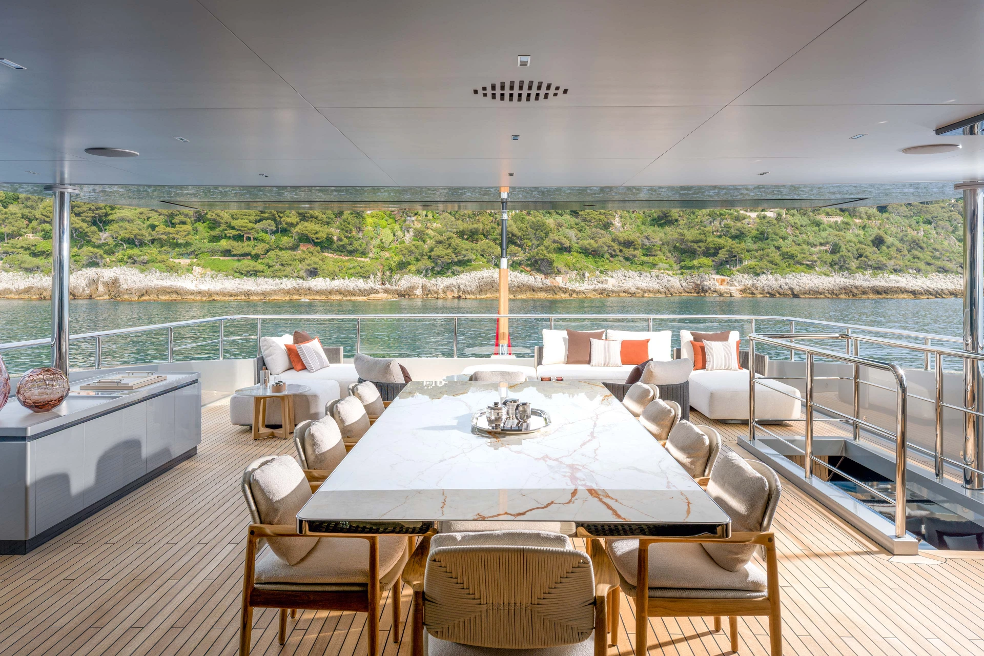 A long marble dining table on a covered aft deck, sofas behind it and a wooded shoreline past the rail