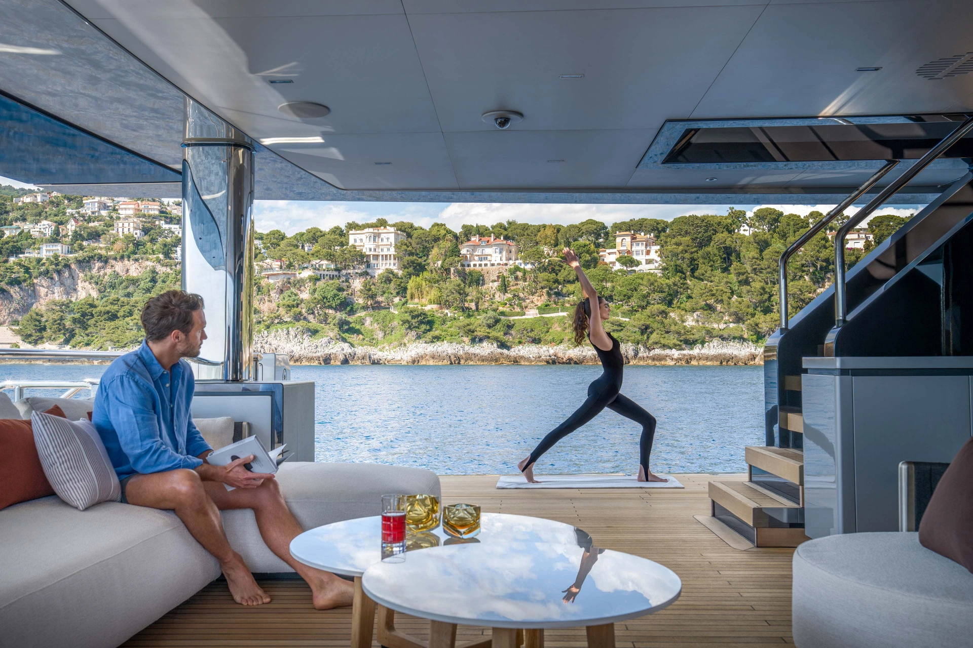 A photograph looking aft through the main deck saloon to the open deck, one guest reading and another doing yoga, a wooded headland beyond
