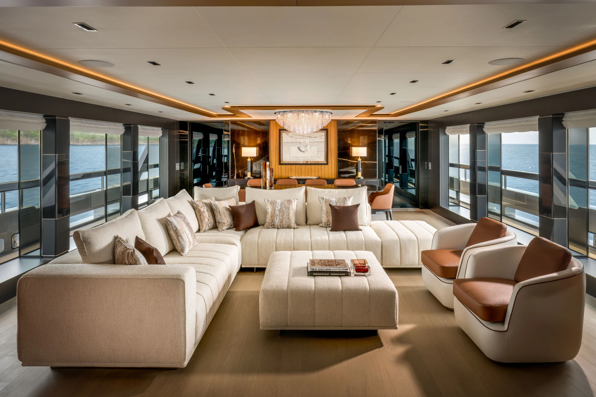 A photograph of the main saloon, pale sofas around a large ottoman with glazing down both sides and a dining area beyond