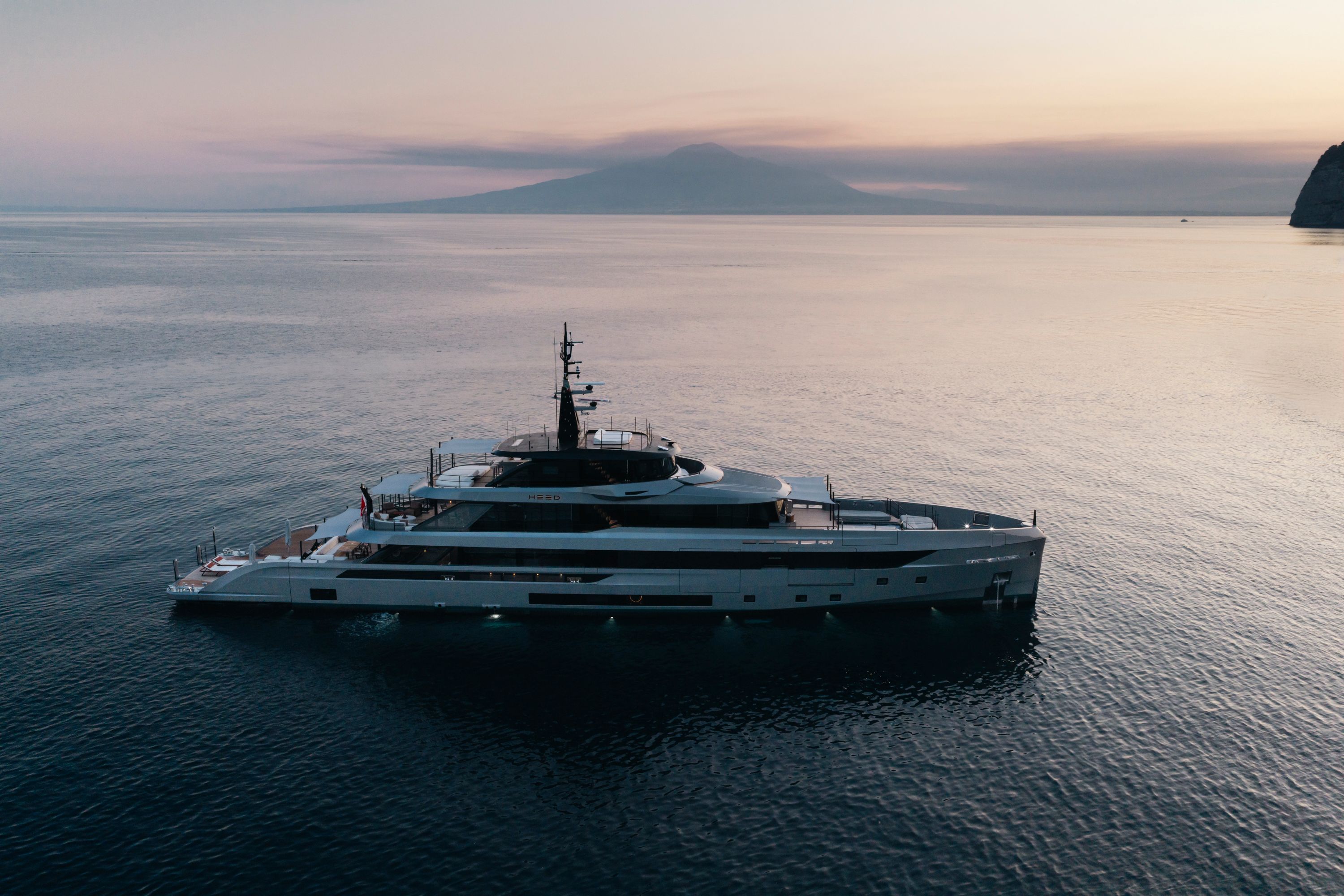 HEED seen from the air at sunset, her aft decks, sundeck and tender stowage laid out below, Vesuvius across the bay