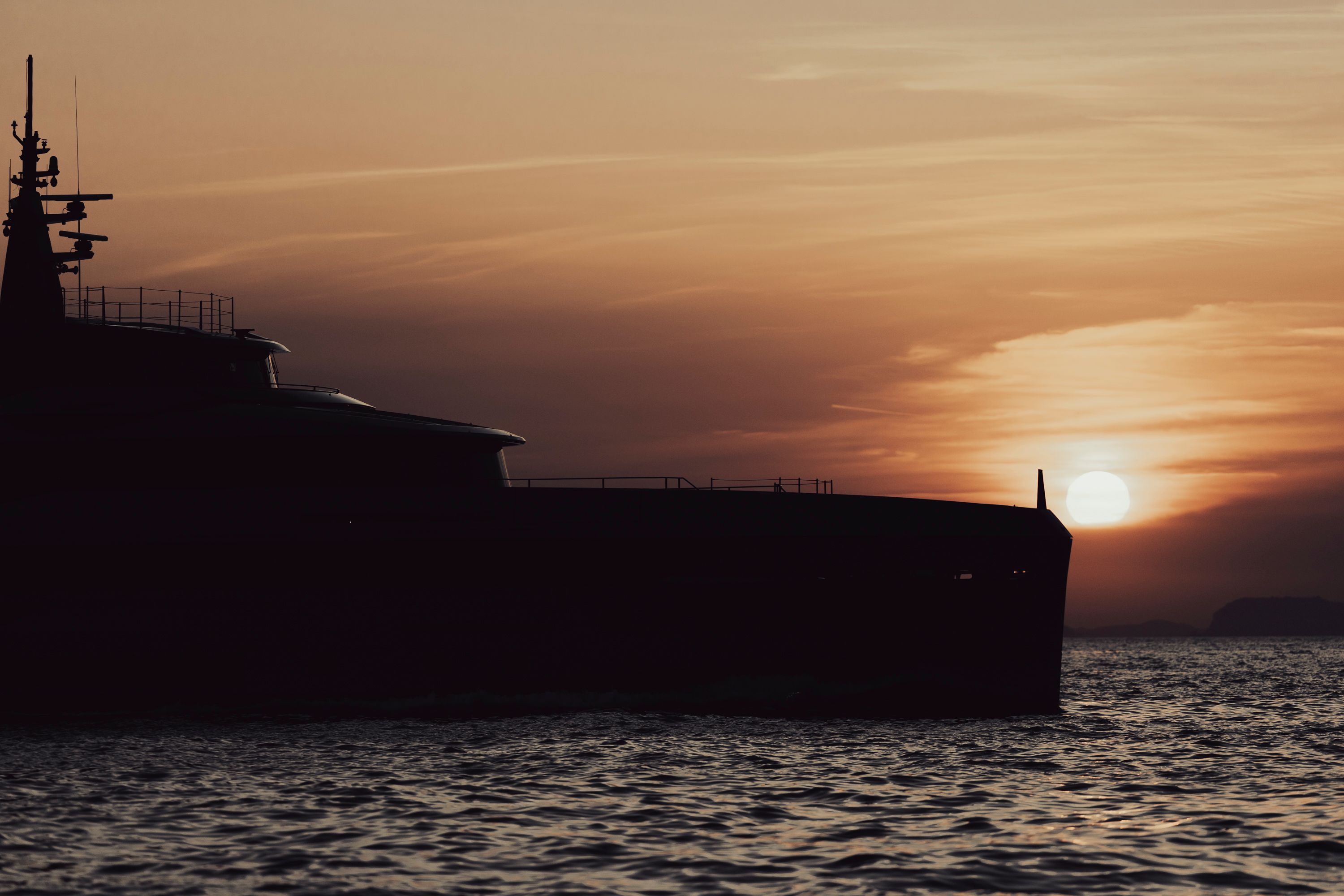 The bow half of HEED in silhouette against a low orange sun, her sheer line running flat to a vertical stem
