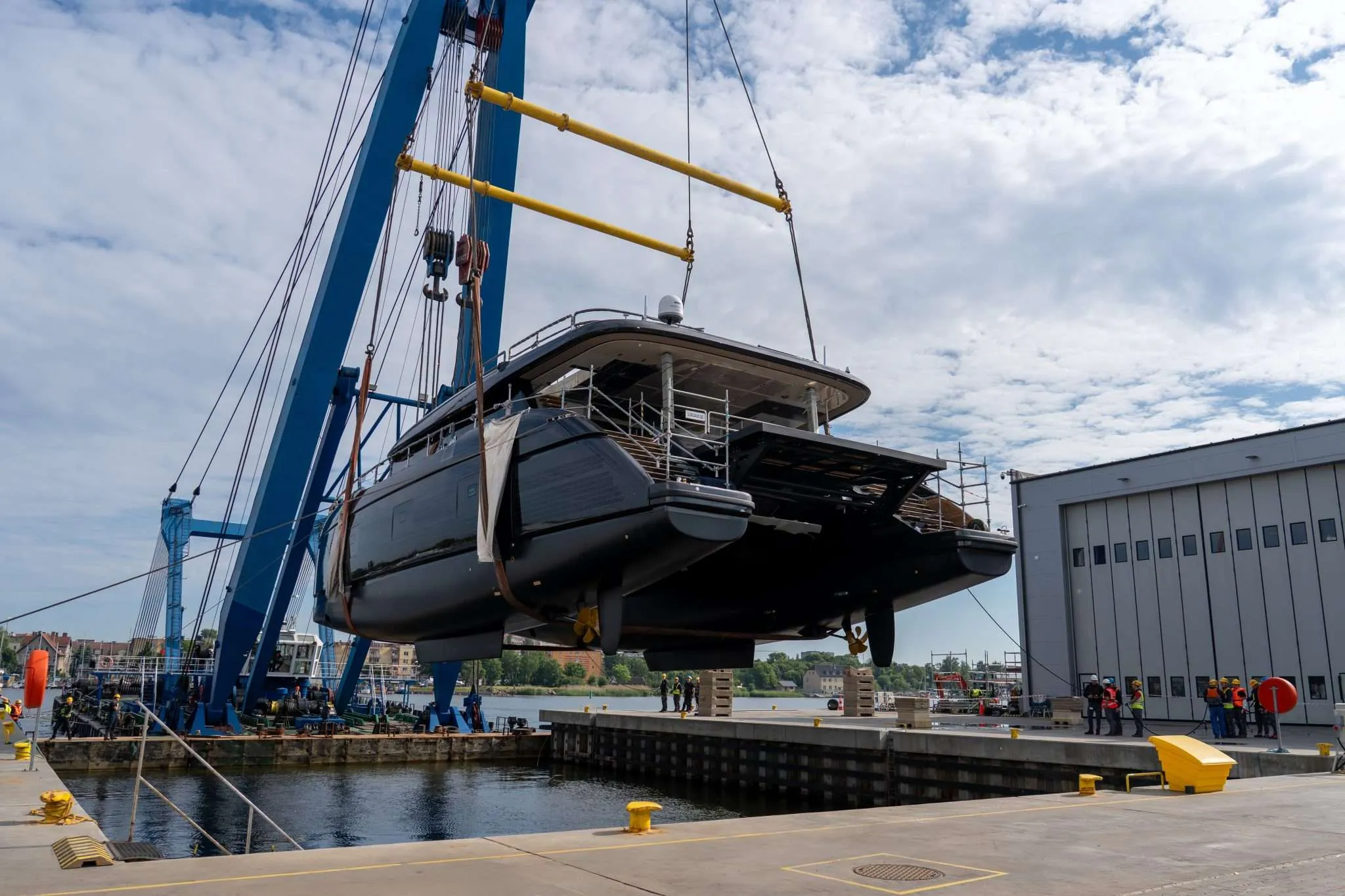 The first Sunreef 100 Eco slung under a blue travel crane above the water, her twin dark hulls and bronze propellers clear of the quay