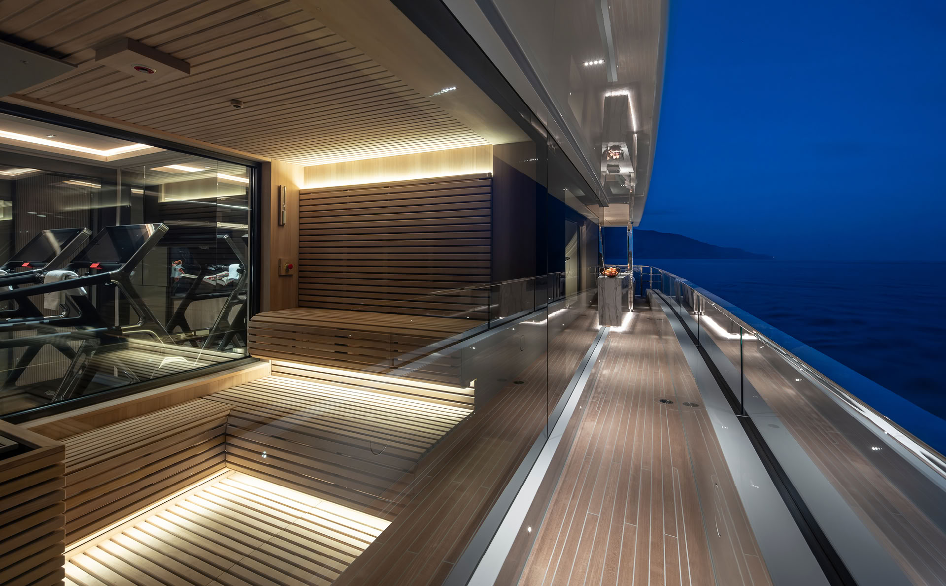 A photograph at dusk of the gym behind glass on the upper deck, a slatted timber bench outside it and the side deck running aft with the coast beyond