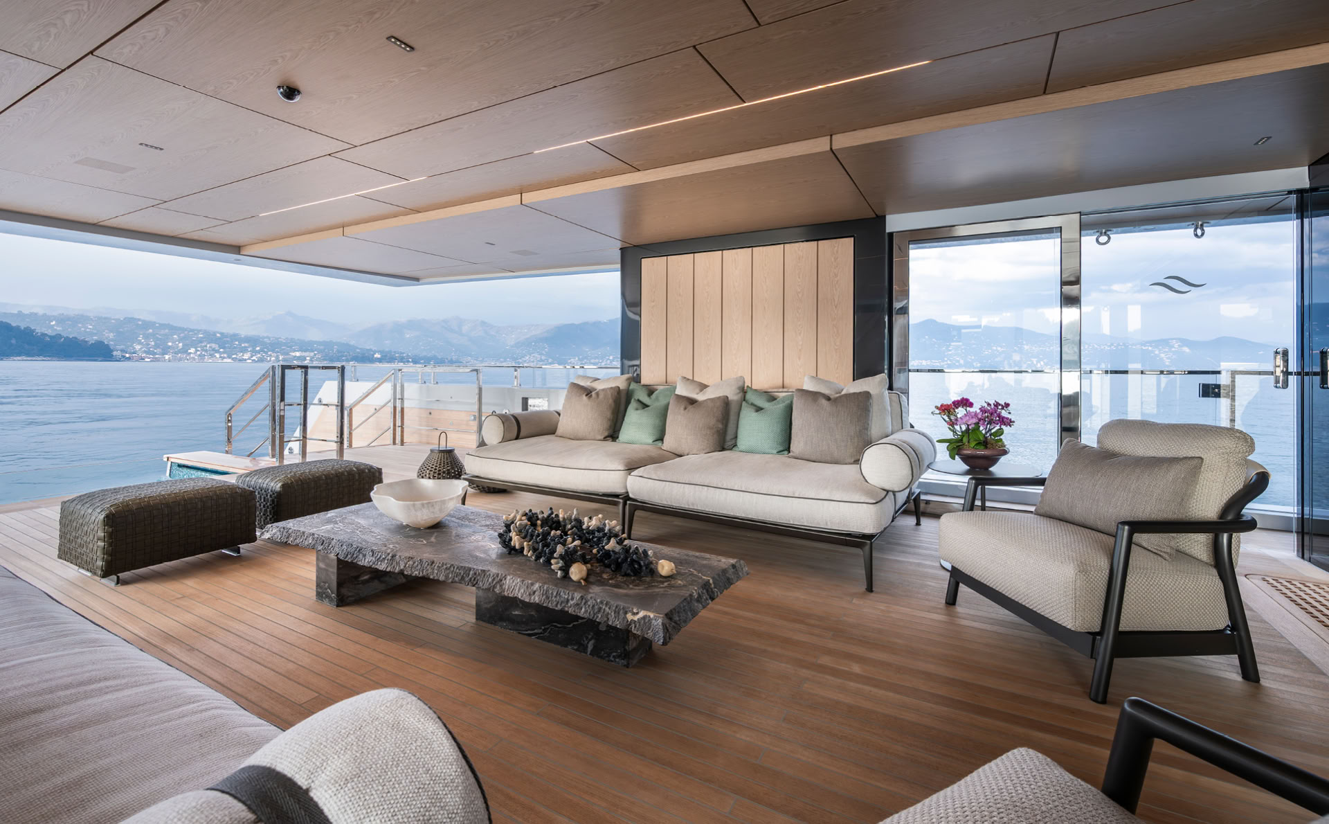A photograph of an open-sided deck lounge with pale sofas, a slab stone table and a timber ceiling, the pool and the sea beyond