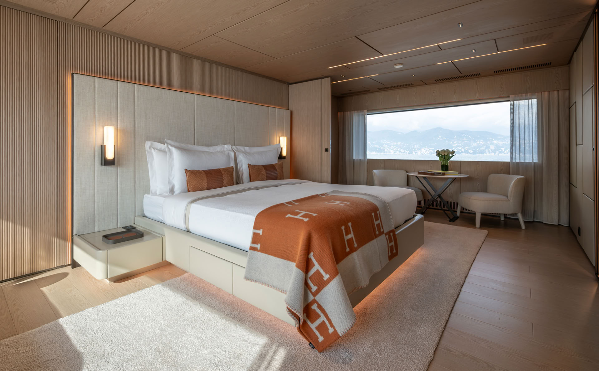 A photograph of a cabin with a double bed against a fabric headboard, wall lights either side and a wide window looking out to a hillside town