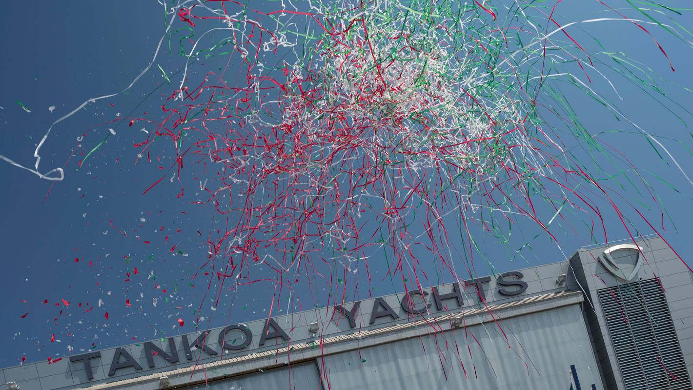 A photograph of red, white and green streamers bursting over the Tankoa Yachts shed against a blue sky